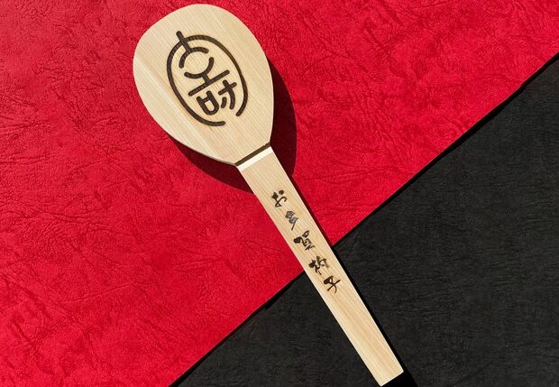 多賀大社の授与品である大きな木製の「お多賀杓子」と、数膳の「多賀長寿はし」が木製の台に並べられている様子。