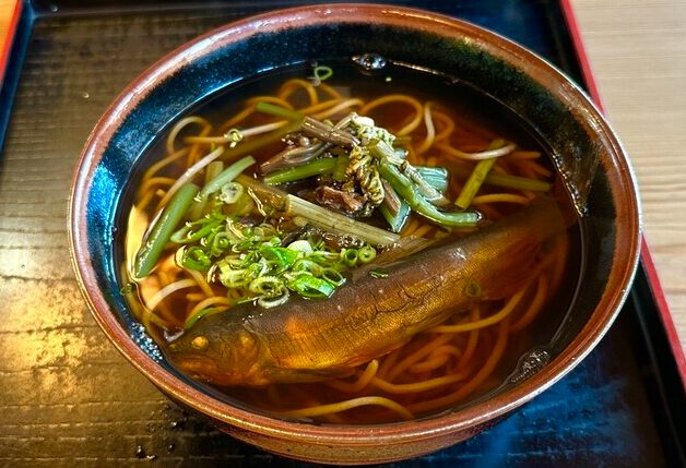 湯気が立つ温かい「寿命そば」の器。香ばしく焼かれた鮎が一匹乗っており、箸で蕎麦を持ち上げている様子。背景は落ち着いた和風の店内。