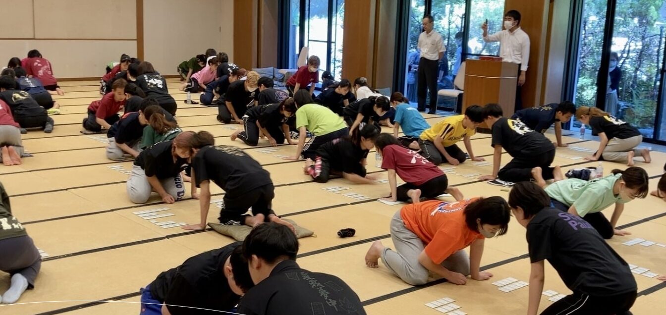 畳の大広間で行われている競技かるたの試合風景。袴姿の高校生たちが真剣な表情で札を払う瞬間。全国高等学校かるた選手権大会（かるたの甲子園）の熱気が伝わる。