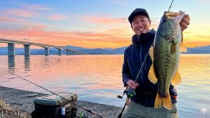 美しい夕焼けに染まる琵琶湖岸で、釣り上げた大きなブラックバスを誇らしげに掲げて笑顔を見せる男性釣り人。背景には琵琶湖大橋のシルエットと広大な湖面が広がり、手前にはタックルボックスと釣り竿が置かれている、初心者向け釣りガイド記事のアイキャッチ画像。