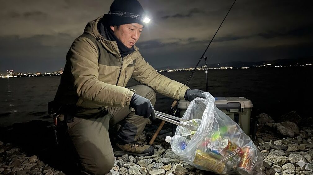 夜の琵琶湖岸で、環境保護のために自主的にゴミ拾いを行っている釣り人の様子。マナー遵守の啓発。