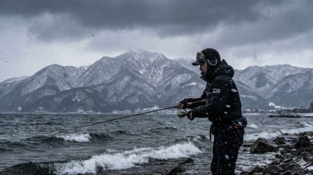 雪が舞う冬の琵琶湖で防寒着を着てバス釣りをする釣り人。比良山系の雪景色と厳しい寒さが伝わる様子。