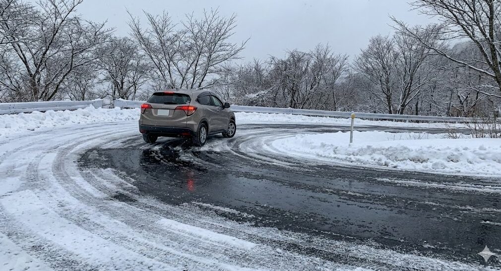比叡山ドライブウェイの冬の路面状況。雪とブラックアイスバーンが混在する危険なカーブを、スタッドレスタイヤ装着車が慎重に走行している様子。